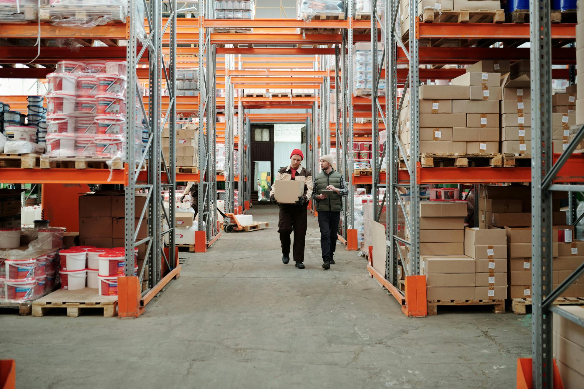 Dos trabajadores manipulan un paquete en un amplio almacén rodeado de estanterías repletas de cajas y productos.